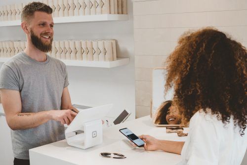 Homem atrás de caixa de loja atendendo uma cliente sorrindo, representando o relacionamento com o cliente.